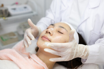 Naklejka premium Woman relaxes during facial treatment. Professional hands gently massage her forehead. Eyes closed, she appears serene and calm. Soft lighting enhances tranquil spa atmosphere 