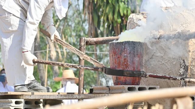 Blacksmith holding a hot crucible by the iron pliers from furnace and pouring the melting gold into the statue block. The process to making of metal statue in local Thailand traditional.