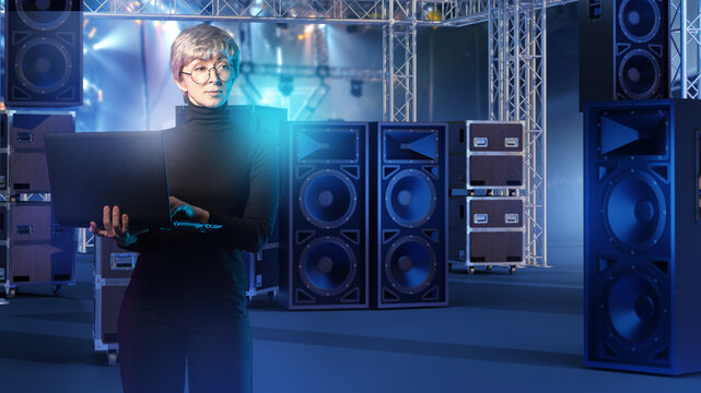 Event production, Audio engineering. Technician configures concert sound system with laptop among stage speakers and truss structures.