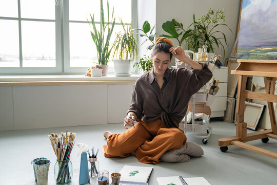 Calm woman painter getting inspiration while creating new masterpiece, sitting on floor drawing with watercolor, female painter painting picture with aquarelle, immersed in thinking process.