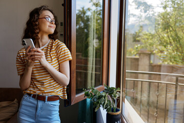 Fototapeta premium Young woman stands by an open window at home, gazing outside while holding a smartphone, wearing a striped yellow shirt and jeans, with a plant on the sill and bright natural light.