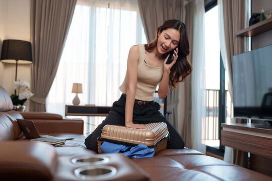 Woman struggling to close overpacked suitcase talking on phone