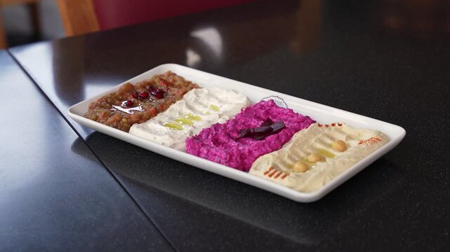 A white platter of assorted Middle Eastern dips including beetroot hummus and baba ghanoush served on a dark restaurant table in natural daylight during a lunch meal.