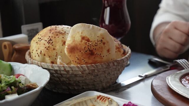 Close up of a man's hand picking up a piece of puffed pita bread from a wicker basket. Served with hummus and salad at a Turkish restaurant during lunch.