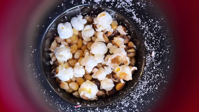 Top-down close up shot of corn kernels popping inside a machine with salt granules. Bright indoor lighting highlights the transition from yellow seeds to white fluffy popcorn during preparation.
