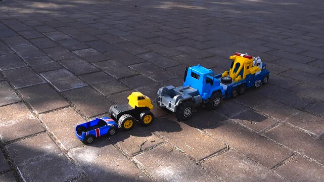 Colorful toy trucks and a race car forming a convoy on a paved ground outdoors during a sunny day. A blue carrier is towing another truck, showcasing a childhood play scene with vehicles