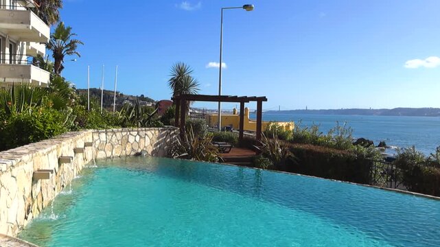 Stunning view of a beautiful infinity pool at a luxury resort, with water jets flowing and overlooking the Tagus river with the 25 de Abril Bridge in the background on a sunny day