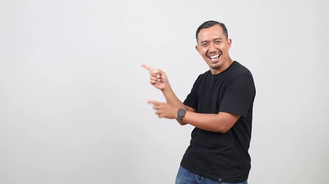 Asian man looks with interest and amazement aside indicates at copy space shows good deal recommends product isolated on white studio wall