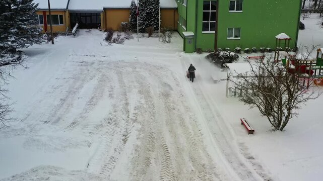Person pushing a snow blower clearing a white winter yard by residential buildings