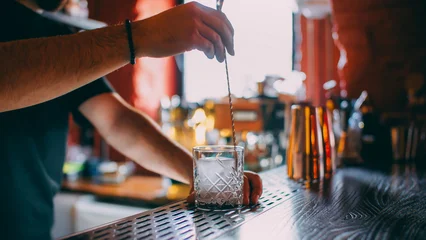 Cocktail making. Ice cube mixing. Unrecognizable barman hands stirring alcohol drink with professional spoon in transparent glass on bar counter. © golubovy