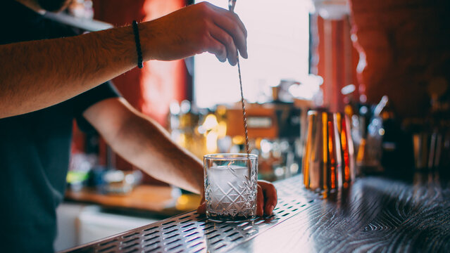 Cocktail making. Ice cube mixing. Unrecognizable barman hands stirring alcohol drink with professional spoon in transparent glass on bar counter.