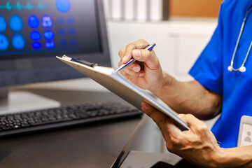 Fototapeta premium Doctor or nurse in blue scrubs writing on clipboard beside computer monitor with medical imaging graphics in clinical office, close-up hands, healthcare documentation concept.