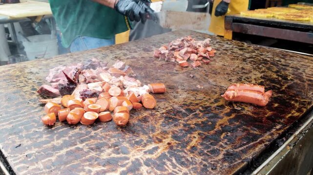 Chef griddling pork meat and sausages at dinner party outside - Close up male hands cooking meat on a flat-top grill for family in restaurant backyard - Food and summer activities