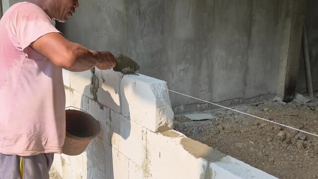 Close-up footage of a skilled construction worker carefully placing hebel (autoclaved aerated concrete) blocks and pouring wet cement mortar between them during a building construction process