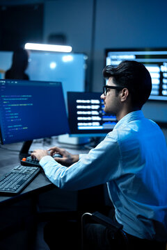 Young male software engineer writing program code on multiple desktop screens while his colleague working in the background in a modern office.