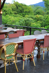 Fototapeta premium Empty bistro tables and woven chairs sit quietly on a balcony, waiting for guests to enjoy a meal overlooking the lush tropical forest and distant mountains.