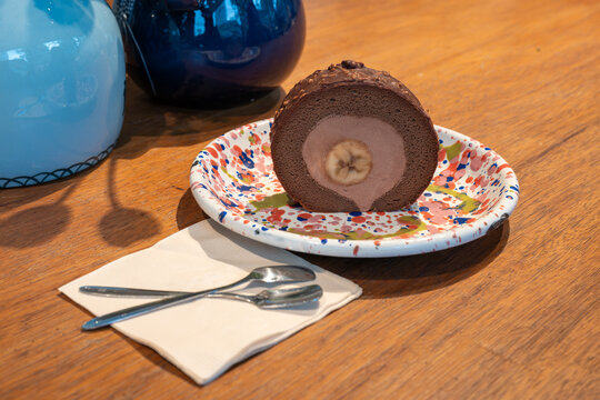 A slice of chocolate cake with a banana filling sits on a colorful plate, surrounded by blue and white decorative jars on a wooden table. The focus is on the cake, highlighting its rich texture and