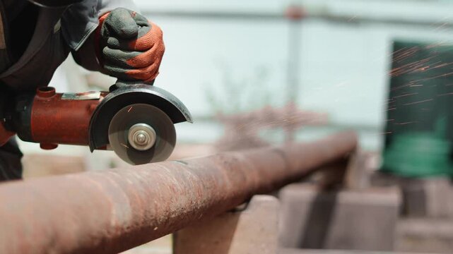 Industrial worker cuts rusted steel pipe with electric grinder during outdoor construction process. Flying sparks, protective gloves highlight precision, safety standards and heavy metal fabrication