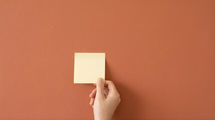 Obraz premium Hand holding a blank yellow sticky note against a minimalist warm terracotta wall background for copy space