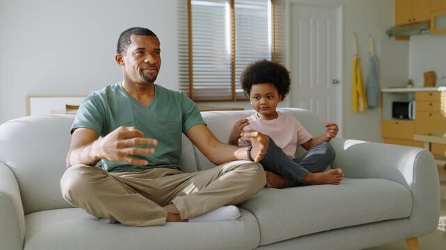 African father and son meditating together on sofa at home.