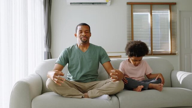 African father and son meditating together on sofa at home.