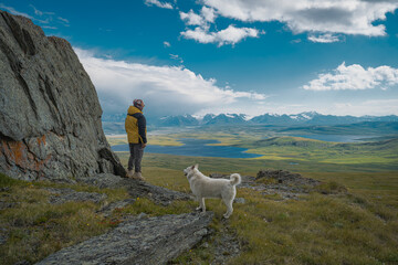 Man hiker and white dog standing on rock looking at scenic mountain lakes. Male traveler with husky exploring wilderness valley, outdoor travel adventure, green landscape and lifestyle © ANDREY