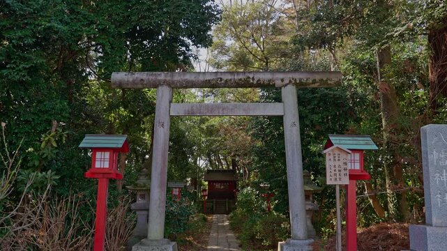 A weathered stone torii gate flanked by traditional red lanterns stands at the entrance of a tranquil Shinto shrine, framed by dense green trees and leading to a vermilion sanctuary beyond.