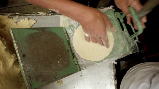 Close view of worker shaping fresh corn tortilla dough using traditional metal tortilla press during authentic Mexican food preparation process