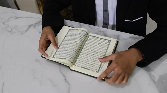 Close up video of man in black suit reading and pointing Al Quran holy book open on white marble table
