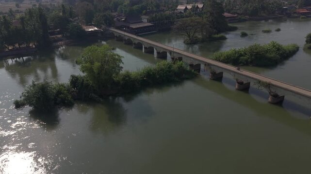 Slow cinematic tilt-up aerial of the Don Det&ndash;Don Khon Bridge over the Mekong River in Laos, with atmospheric haze and distant hills in the background.