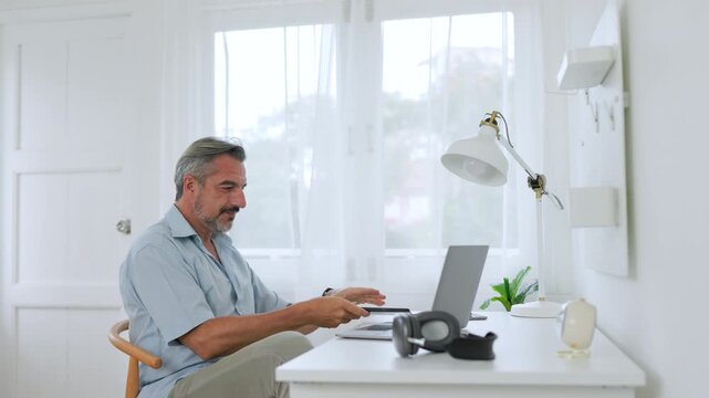 Mature businessman using smartphone while working on laptop in modern home office.