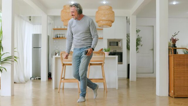 Happy mature man dancing with wireless headphones and smartphone in modern home kitchen.