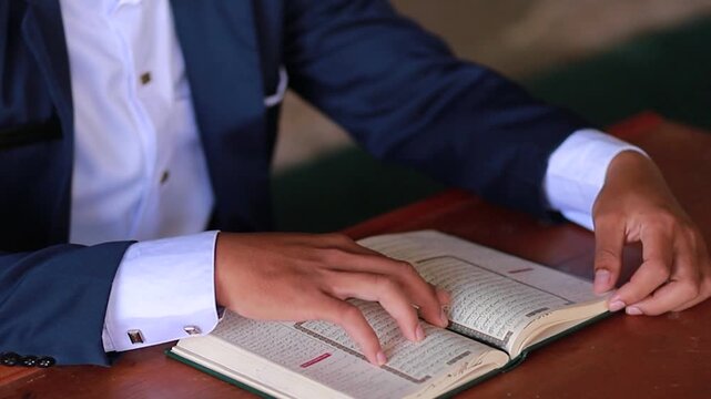 Close up video of man in formal suit reading and reciting Al Quran holy book on wooden table with both hands touching pages