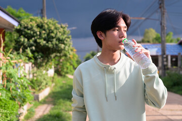 Fototapeta premium Young Thai man from Isan drinking bottled water looking away outdoors