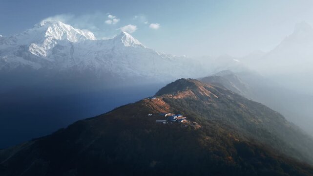 Aerial drone footage of a forested mountain ridge with a small lodge settlement, set against a backdrop of misty Himalayan peaks rising behind veils of atmospheric haze in Nepal.