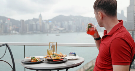 Obraz premium Man in red polo shirt drinking cocktail at rooftop bar facing Hong Kong skyline and Victoria Harbour. luxury lifestyle, travel destination, urban dining, vacation, metropolis, asian tourism.