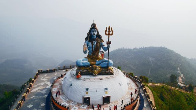 Aerial drone shot of the massive Pumdikot Shiva Statue in Pokhara, Nepal, showing the blue deity seated atop a white dome platform with the misty hills and valleys of the Pokhara region in the backgro
