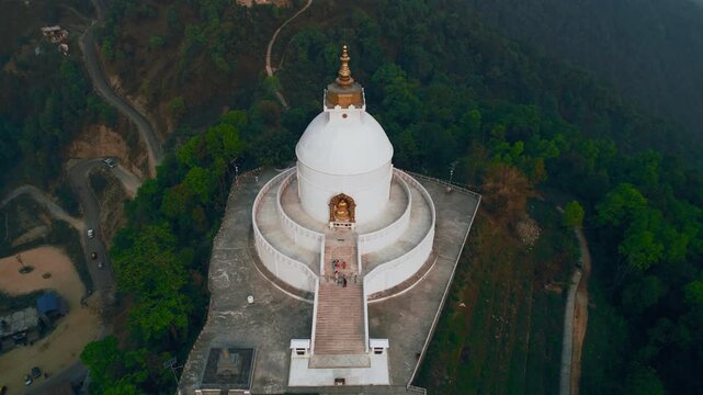 An aerial drone shot captures the gleaming white World Peace Pagoda crowned with a golden spire rising above the forested hills of Nepal.