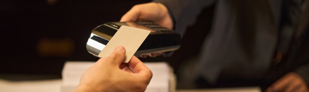 Close up of young adult customer holding bank card near payment terminal, making contactless purchase at checkout. Ideal for retail payment, fintech, consumer spending marketing, header