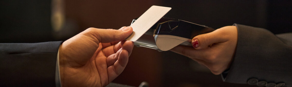 Close up of business traveler handing payment card to receptionist processing transaction on card terminal. Supporting hotel check in, corporate travel, hospitality marketing, header