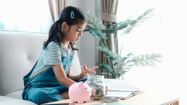 A young Asian girl sitting on a grey sofa at home, counting on her fingers to calculate her savings with a pink piggy bank and a coin jar nearby.