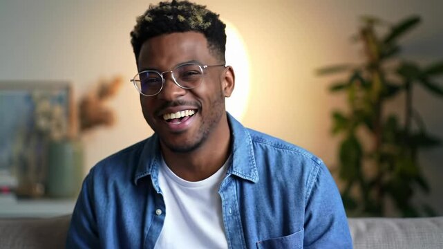 A young afro-american man with curly hair, stylish round glasses, and a blue denim shirt laughs, talks, and gestures in a modern indoor setting with soft bokeh. online communication concept