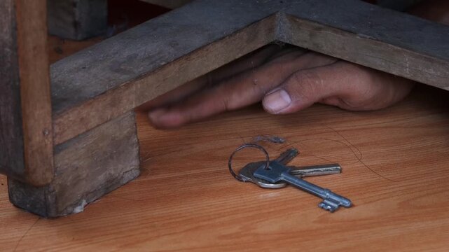 Close up of Asian man hand reaching hidden house keys under wooden table indoors. Secret spare key storage concept for home access, security, emergency backup, and property safety.