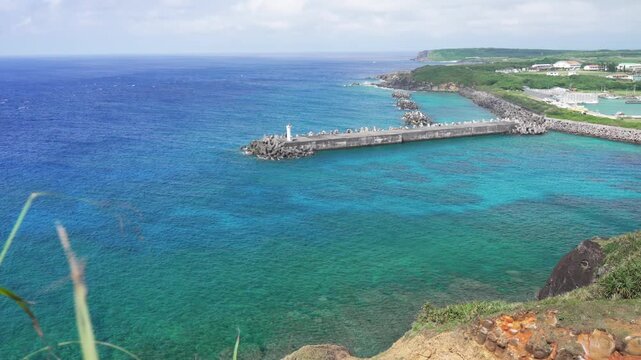 Scenic view of a port and beautiful blue ocean with coral reef from Irizaki Lighthouse observatory in Yonaguni Island, Japan's westernmost point. Green grass swaying in the wind.