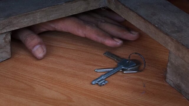 Close up of Asian man hand reaching hidden house keys under wooden table indoors. Secret spare key storage concept for home access, security, emergency backup, and property safety.