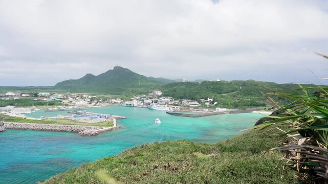 A sightseeing boat slowly turning and entering a harbor surrounded by emerald green sea and hills in Yonaguni Island, Okinawa, Japan.
