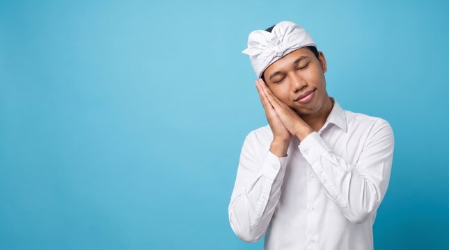 Tired young Balinese man wearing Indonesian traditional udeng falling asleep isolated on blue background