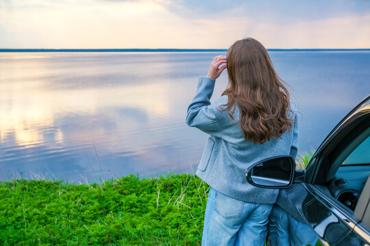 A young woman in casual clothes near the car enjoys the scenery of the lake at an overcast sunset. travel, adventure, pause during a trip