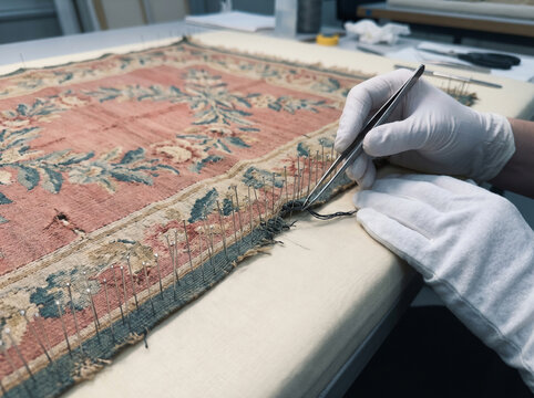 Antique Aubusson flatweave fragment pinned to padded restoration table with conservator tweezers lifting broken weft thread