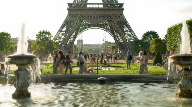 Wide video of iconic Eiffel Tower over Trocad&eacute;ro Gardens. Ornate fountains spray water into a pond, diverse people stroll on sunlit paths, relaxing on green grass. Serene travel concept